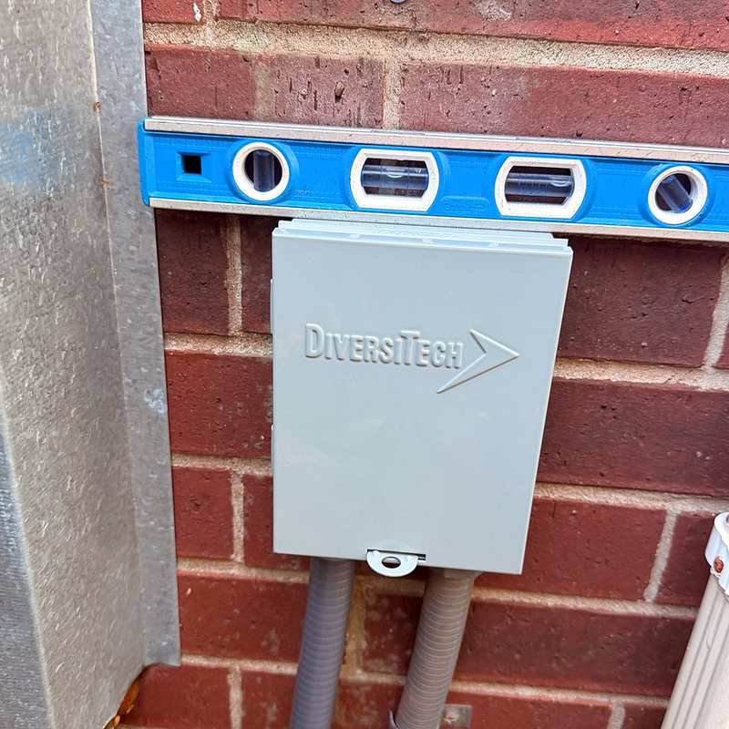Close-up of a DiversiTech electrical disconnect box mounted level on a brick wall beside a condenser