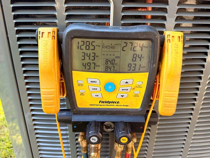 Close-up of Roo-D's Fieldpiece manifold gauge clipped to an outdoor condenser, showing live refrigerant readings